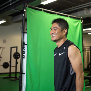 Close-up of Shinji Kawashima, smiling and standing in front of a green screen. He's wearing a Nike jersey. The setting is an indoor gym.