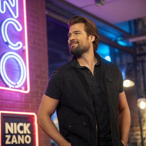 Nick Zano, bearded actor, dressed casually in a black vest. On set of a TV show. Bright neon sign with studio logo in background.