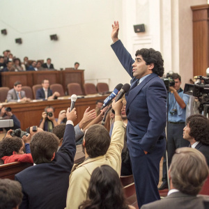 Diego Maradona, in a navy blue suit, at a press conference. Crowd of journalists with raised hands