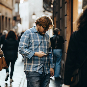 Ed Sheeran, a white male, wearing a blue plaid shirt and jeans, checking his phone amidst a crowded street.