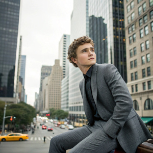 Callan McAuliffe in a gray suit with curly hair, sitting amidst a bustling cityscape, skyscrapers towering in the background.
