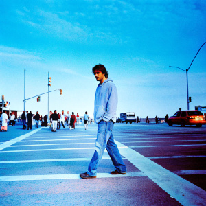 Robert Pattinson in a candid shot. Casual outfit, jeans, and a light blue hoodie. Crowded city street backdrop.