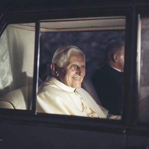 Pope Benedict XVI smiling through a car window. Dark, shadowy backdrop. Solemn procession