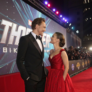 Tom Hiddleston and Natalie Portman at a glamorous movie premiere. They are standing in front of a giant LED screen displaying the film's promotional content.