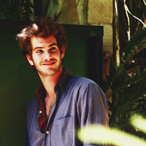 Andrew Garfield, actor, with a charming smile, standing in front of a green screen. His disheveled hair and slight stubble make him look ruggedly handsome. Wearing a light blue button-up shirt. Background blurred.