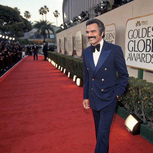Burt Reynolds, in a navy blazer and matching pants, attends the Golden Globe Awards. Smiling, his iconic mustache stands out. Event hall with red carpet and flashing cameras