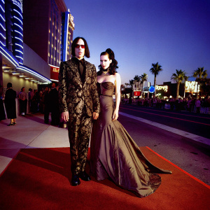 Marilyn Manson and Dita Von Teese at a music awards show. Manson in a black and gold embroidered suit, Dita in a brown, floor-length gown. Neon lights and crowd in background