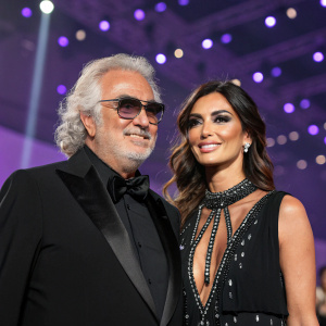Flavio Briatore and Elisabetta Gregoraci at a glamorous gala. Flavio in a black tuxedo, Elisabetta in a dramatic black dress with detailed embellishments. Purple backdrop with sparkling lights