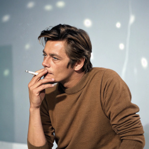 Alain Delon, Caucasian actor, in a casual brown sweater, smoking a cigarette. Shot in a modern, minimalist studio setting. The background is a soft pastel grey with subtle highlights.