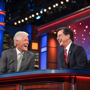 Bill Clinton and Stephen Colbert laughing together on The Late Show set, bright studio lights.
