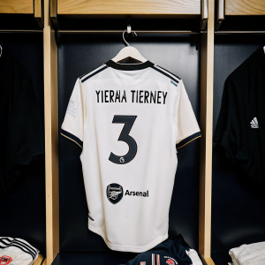 Kieran Tierney's Arsenal jersey, number 3, hanging in a bustling locker room. Wood panels, metal hooks, and various team patches surround the jersey.
