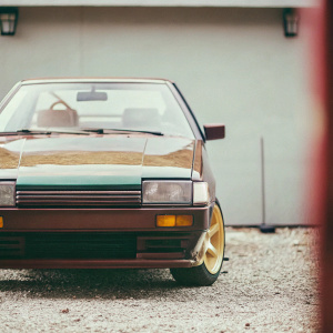 A dark green AE86 Trueno parked in an open garage. Golden rims and a lowered stance giving it a fierce look. Inside is a small, cartoonish toy, adding a playful touch. The neutral grey walls provide a backdrop for this custom creation.