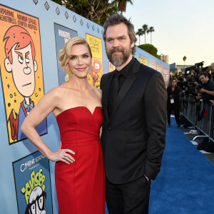Rhea Seehorn and Graham Larson at the Critics' Choice Television Awards. Rhea wearing a stunning red off-the-shoulder gown, Graham in a sleek black suit. They stand against a vibrant backdrop featuring playful caricatures and cartoon-like artwork. The event is bustling with celebrities and paparazzi.