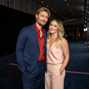 Chad Michael Murray and Hilary Duff, posing for an interview at a star-studded gala. Chad in a navy suit with a vibrant red shirt underneath. Hilary dons a soft peach top. They stand on a dark, textured carpet with a dimly lit backdrop.