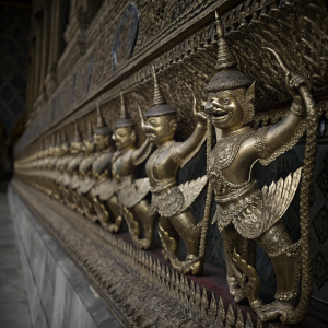 Golden Garuda statues lining the walls of the Grand Palace's Emerald Buddha Temple. Intricate Thai mythical creatures, detailed in vibrant blue and gold, depicted in dynamic poses against a complex mosaic backdrop.