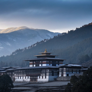 Gantey Monastery, a complex of traditional Tibetan-style buildings nestled in the Phobjika Valley, Bhutan. The main structure, a large wooden building with a distinctive thatched roof, stands out against a backdrop of snow-capped mountains and lush forests.