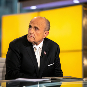 Rudy Giuliani, donning a black suit and white shirt, seated at a news studio desk. Intense facial expression. Bright yellow backdrop.