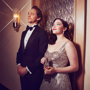 Tom Hiddleston and Kat Dennings attending an awards show. The duo is dressed in formal attire. Tom in a black tuxedo, Kat in a sparkling silver gown. The background features a shimmering backdrop with the event logo.