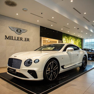 A pristine white Bentley Continental GT V8 parked in a luxurious showroom. Elegant design, exquisite detailing, and the iconic Bentley emblem are prominently displayed. Photographed on a sleek black platform with Miller Motorcars logo visible.