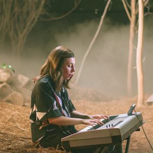 Katelyn Nacon, a young actress, performing at Walker Stalker Con Melbourne 2018. She wears a black shirt with a graphic print and plays a digital piano. Dark, foggy backdrop with a lone tree