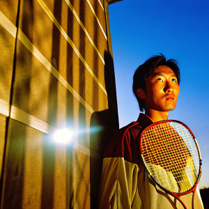 Kento Momota, Japanese badminton player, intense expression, against spotlighted black backdrop. Shining spotlight creating dramatic shadows