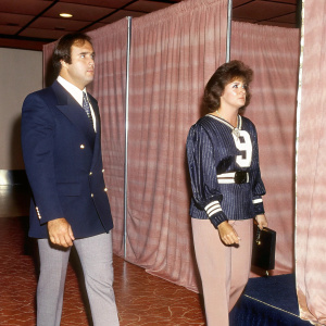 Giorgio Chinaglia and Connie Eruzione at a sports awards gala. Chinaglia, donning a navy blazer with the number 9, stands with a serious demeanor. Eruzione sports a serene expression. The background is a luxurious ballroom filled with cheering fans and flashing cameras.