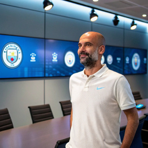 Pep Guardiola, sporting a neat beard, stands in front of a sleek conference room. He wears a crisp white polo shirt. Large screens display team logos.