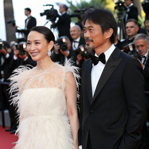 Miki Nakatani and Hiroshi Abe at Cannes Film Festival premiere. Miki in a stunning white feathered dress, Hiroshi in a black tuxedo. Red carpet, flashing cameras, crowd in background.