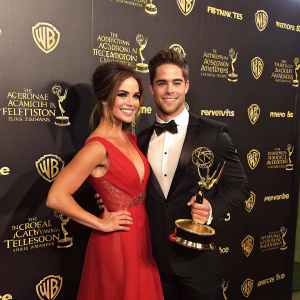 Jillian Murray and Dean Geyer at a glamorous awards ceremony, posing with a backdrop adorned in shining logos.