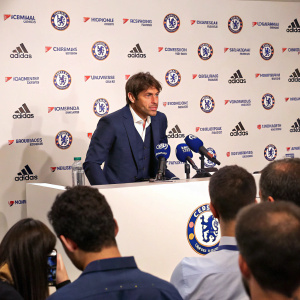 Antonio Conte addressing media in a Chelsea FC press conference. Wearing a navy blue suit. White backdrop with team logo