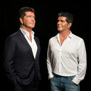 Simon Cowell standing beside a life-sized mannequin resembling a celebrity. Black background, photo