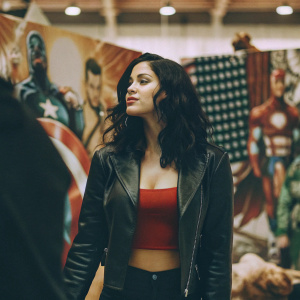 Meta Golding at New York Comic Con. Wearing a sleek black leather jacket over a red crop top. Standing against a vibrant comic book themed backdrop with superheroes