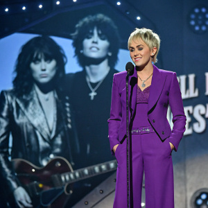 Miley Cyrus, Caucasian, short blonde hair, in purple ensemble. Standing center stage at the 30th Annual Rock and Roll Hall of Fame Induction Ceremony. Surrounded by Joan Jett and the Blackhearts themed backdrop.