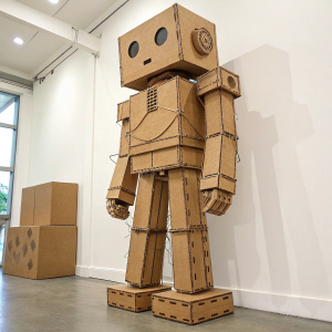 A large cardboard robot sculpture. The torso and head sections look like a giant robot face. The legs are crafted from brown cardboard and string. Simple and creative design displayed on a plain white wall.