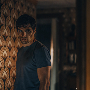 Sam Witwer, portraying Aidan Waite in 'Being Human', stands in a dimly lit room. He has a serious expression, with dark, short hair and blood-like stains on his face. He wears a tight dark blue t-shirt. The background features intricate orange and beige wallpaper.