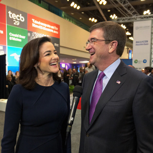 Sheryl Sandberg and Ashton Carter at a tech conference. Sheryl in a dark tailored dress, Ashton in a dark suit with a purple tie. They are candidly chatting near a microphone setup, with conference attendees and event banners visible in the background.