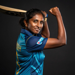 Ranuda Somarathne, a cricket player from Sri Lanka, striking a confident pose. Wearing a blue and teal jersey that fits well. Dark background