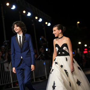 Finn Wolfhard in a navy suit and Millie Bobby Brown in a white and black gown. At the event, they walk towards the crowd. Dark background with flashing lights