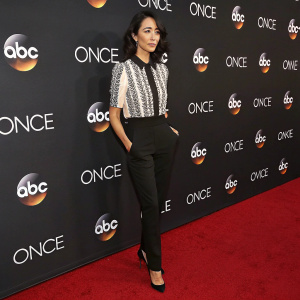Sandrine Holt, actress, stands on red carpet at awards show. Black and white blouse with textured design. Black pants, classic heels. Loose waves hair. Poised and serious expression. ABC and Disney logos visible in background.