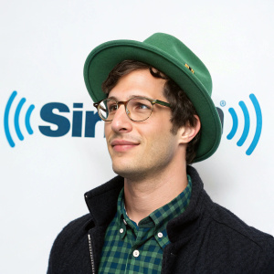 Andy Samberg, fair-skinned, wearing glasses and a green hat, standing in front of the SiriusXM logo. Simple white background, photo