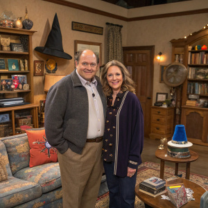 George Costanza and Susan Biddle Ross standing in a cozy living room, amidst magical artifacts and Seinfeld memorabilia.