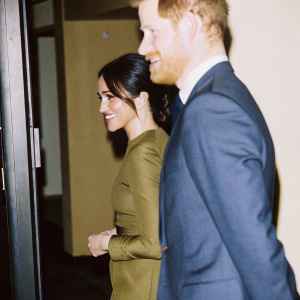 Prince Harry and Meghan Markle, both smiling and standing close. Harry has a beard and navy blue suit. Meghan in green dress. Background blurred.