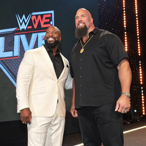 JTG and Big Show, standing side by side at a WWE live event. JTG in a sharp white suit, Big Show in a black shirt. The WWE logo prominently displayed on the backdrop. Big Show with a beard, JTG with a big smile.