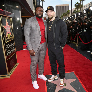 50 Cent and Eminem at a glitzy awards show. Both are standing on a red carpet surrounded by flashing cameras and fans. 50 Cent in a plaid suit, red turtleneck, and white sneakers. Eminem in a black t-shirt and black cap. Backdrop features the Hollywood Walk of Fame.
