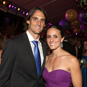 Chris Kluwe and Isabel Kluew, a dashing couple, posing for a picture. Chris in black suit and blue tie, Isabel in a stunning strapless purple gown. Crowded party with festive decorations.