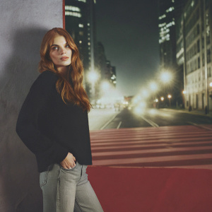 Holland Roden, with long wavy brown hair, standing confidently in a bustling city street. Wearing a black sweater and jeans. Bright city lights in the background.