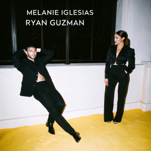 Melanie Iglesias and Ryan Guzman at a glamorous gala. They wear matching black outfits, formal and chic. The backdrop features a sleek metallic logo.