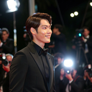 Kim Woo-Bin attending the Cannes Film Festival, dressed in a formal black suit, with a relaxed smile. Bright lights and paparazzi in the background.