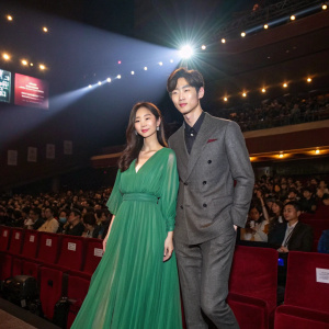 Jin Se-Yeon and Lee Jong-Suk at the 'Doctor Stranger' promotional event. Jin wears a flowing green dress. Lee in sharp grey suit. Crowded auditorium, bright lights
