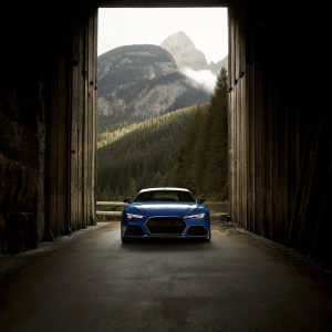 A sleek blue Audi RS7 on a winding road, framed by towering, misty mountains. The car's sharp lines and powerful stance stand out against the dramatic landscape.